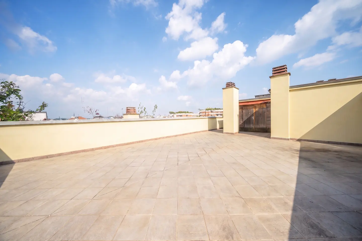 Wide shot of a rooftop terrace with beige tiles, a low wall, and chimney stacks under a blue sky with scattered clouds.