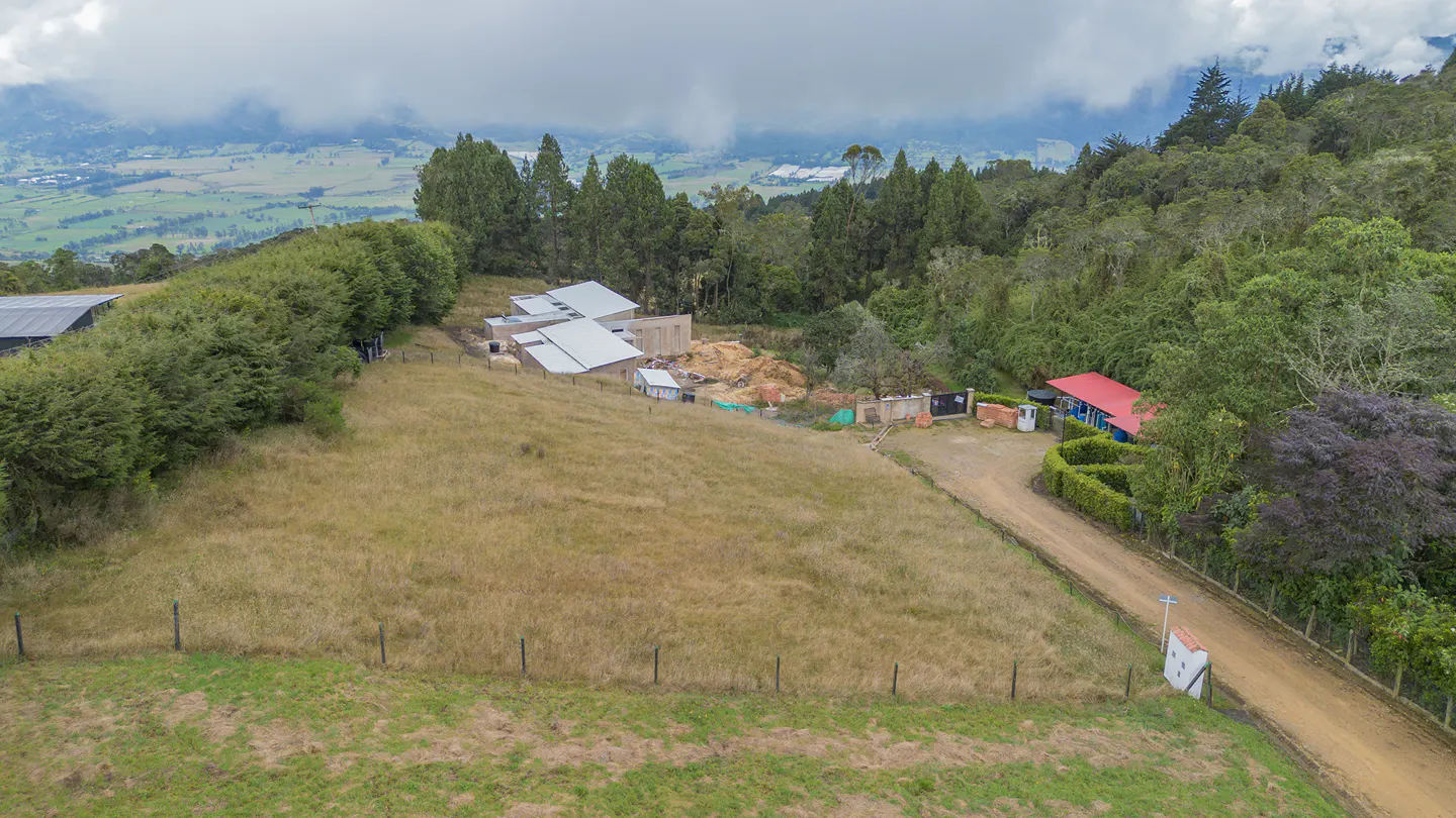 Aerial view of a rural property with a house under construction, a red-roofed house, and a dirt road leading to a small white chapel.