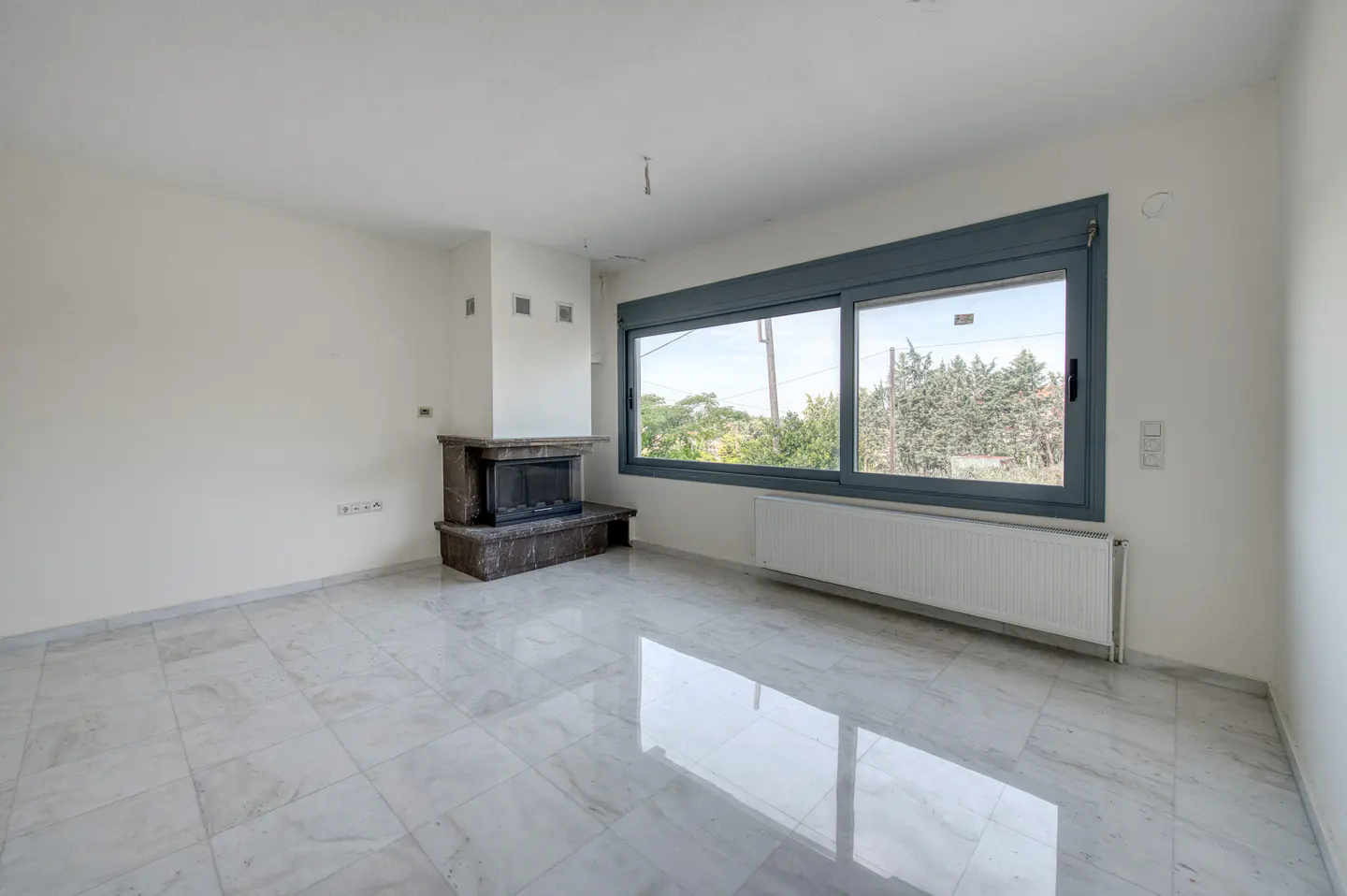 Empty room with white walls, marble floors, a dark stone fireplace, and a large window overlooking trees.