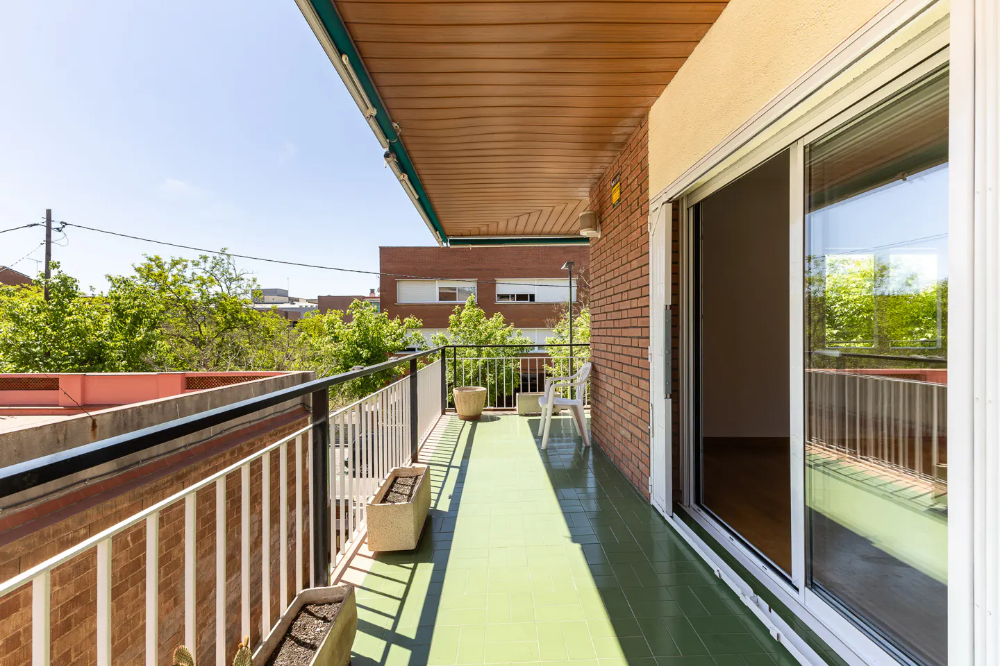 A sunny balcony with green tile floor, white railings, and a sliding glass door. Potted plants line the railing, and trees are visible in the background.