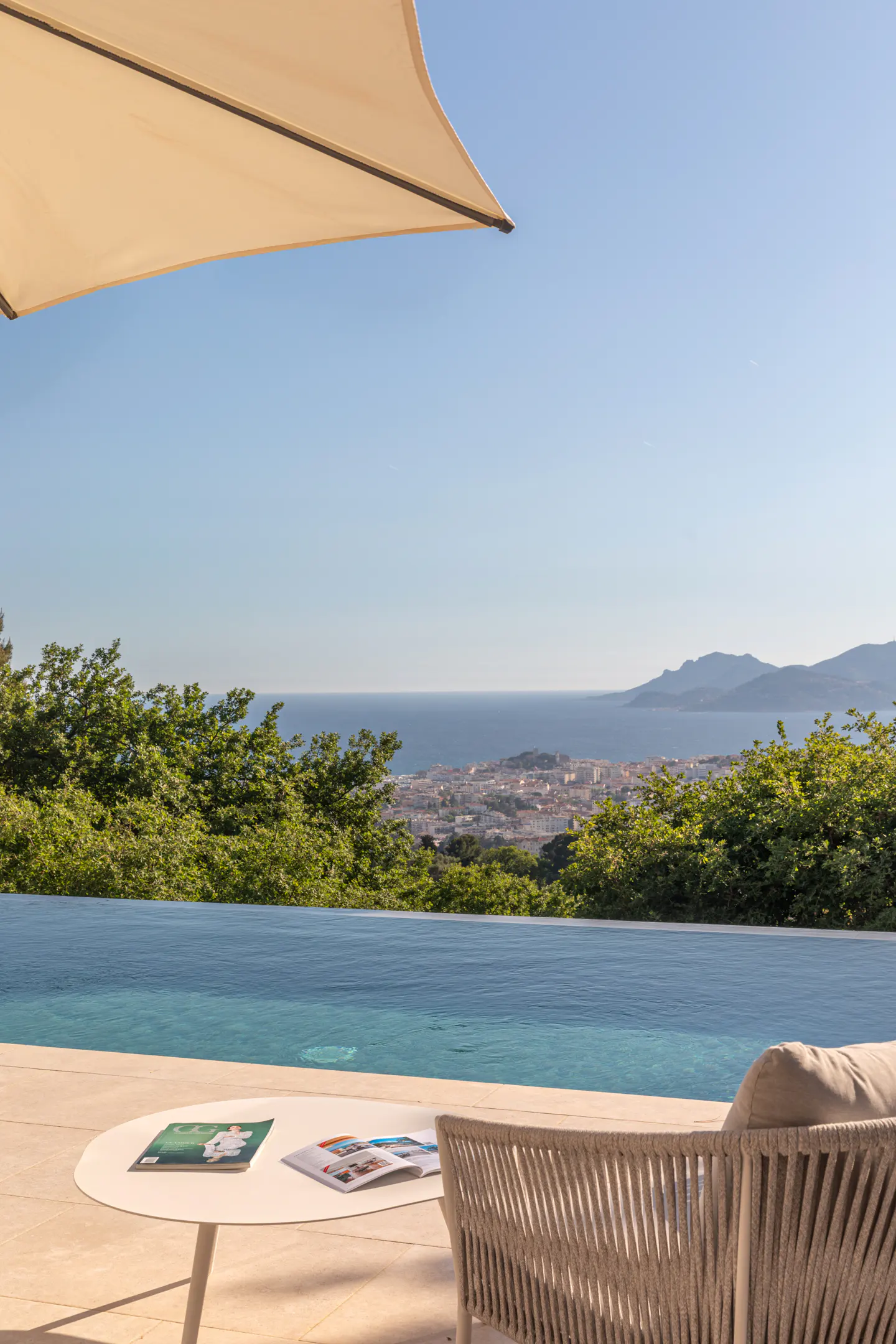 Outdoor patio with infinity pool overlooking the ocean. A white table with magazines and a beige chair are visible.