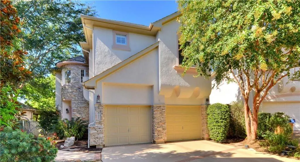 Two-story home with cream stucco, stone accents, and a two-car garage, surrounded by lush green trees on a sunny day.