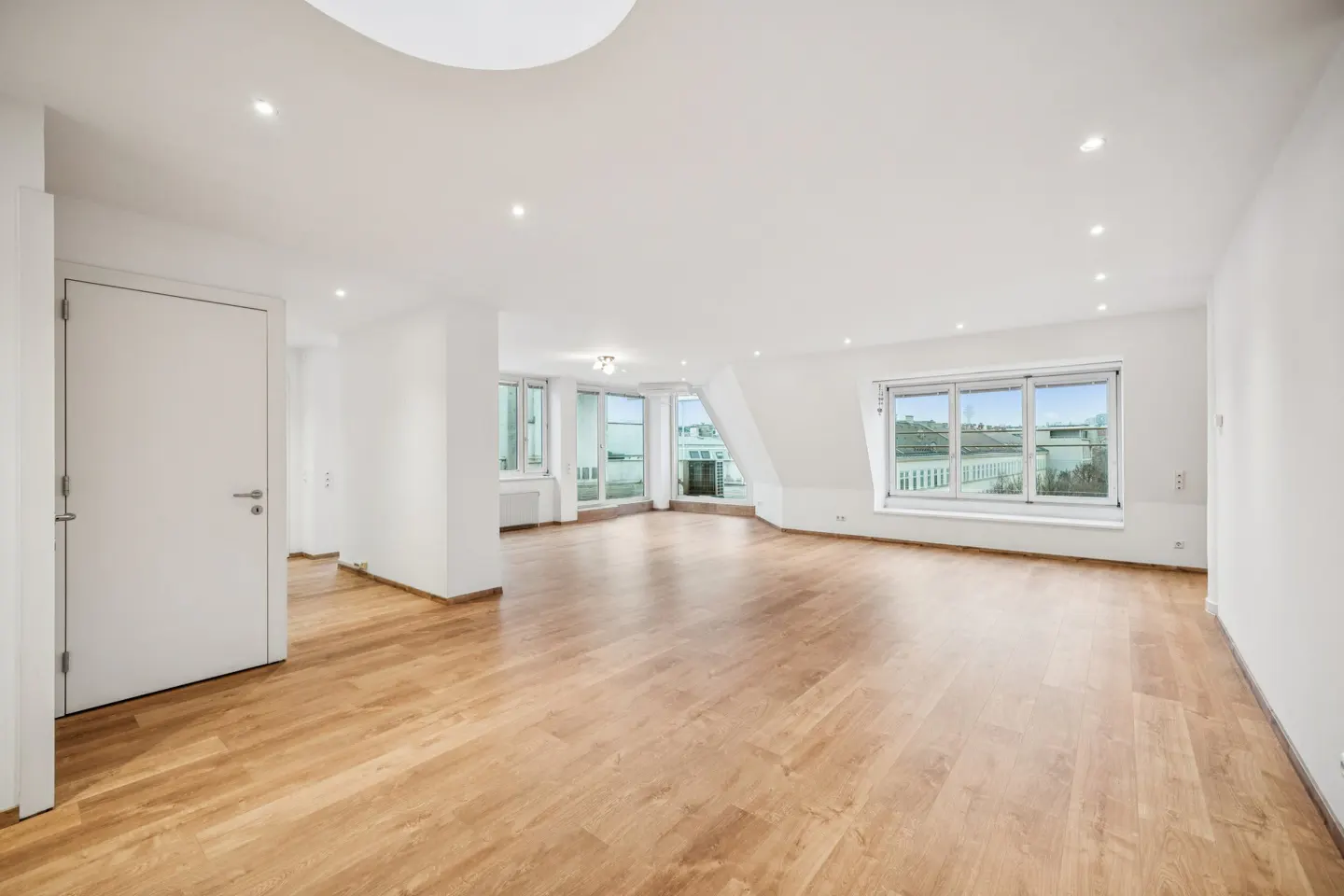 Bright, empty room with hardwood floors, white walls, and recessed lighting. Large windows offer a view of the city.