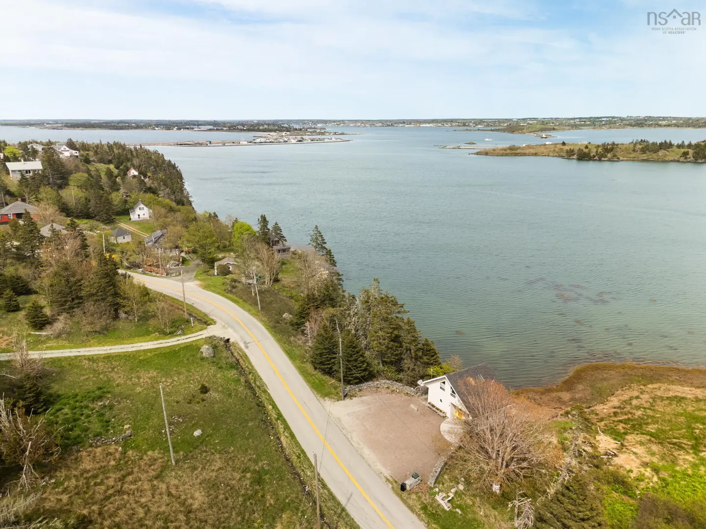 Aerial view of a coastal road winding past houses and trees, leading to a waterfront property with a white house and ocean view.
