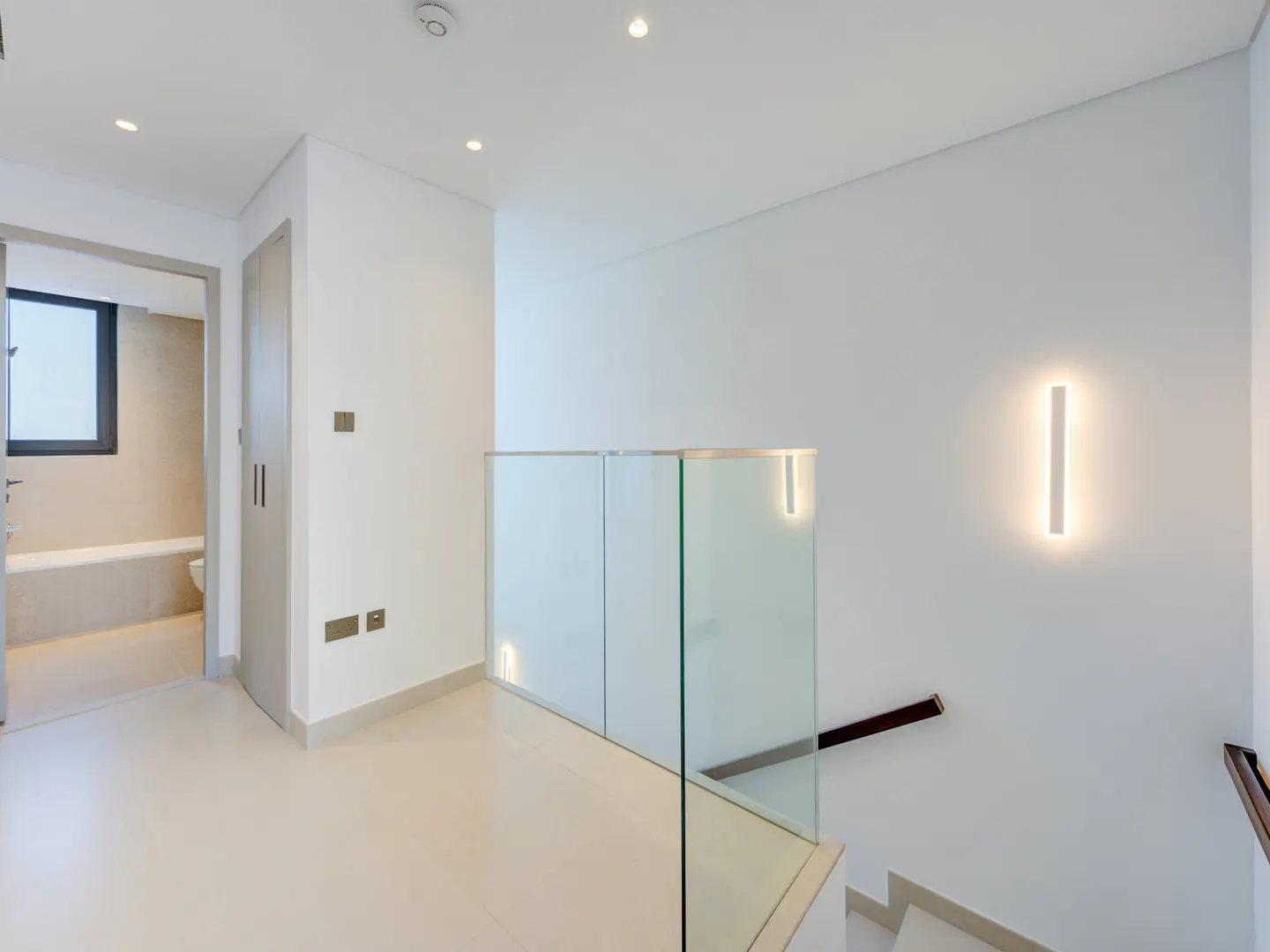 Bright, modern hallway with white walls, light beige floors, and glass railing. A bathroom is visible through an open doorway. A vertical light fixture is on the wall.