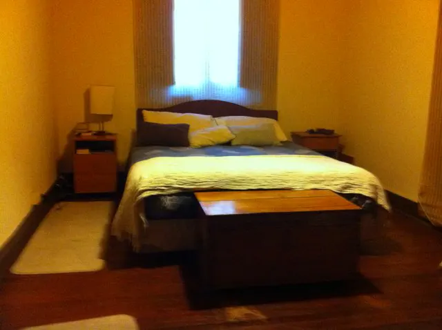 Bedroom with a bed, wooden chest, and nightstands. The walls are yellow, and a bright window is centered behind the bed.