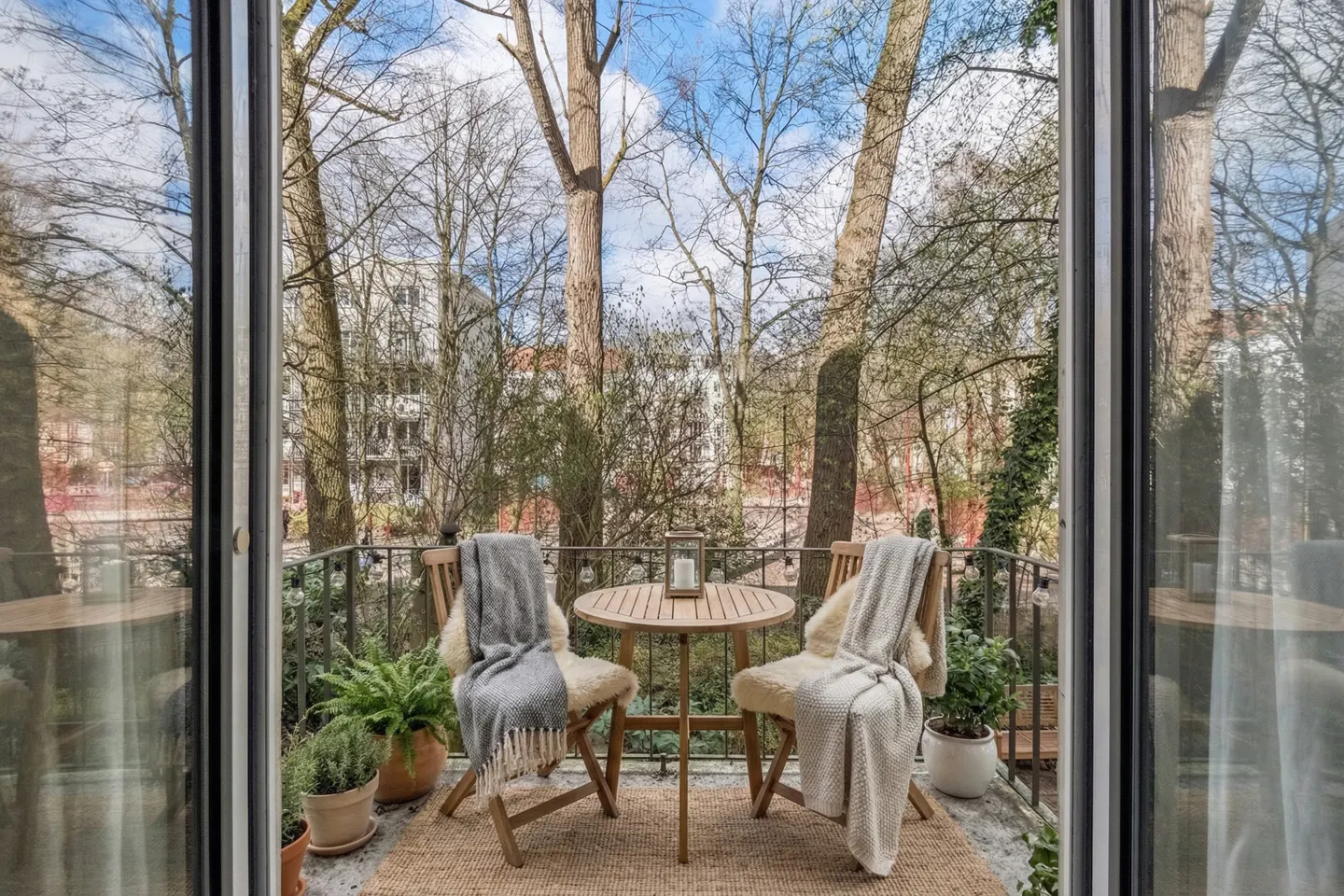 Balcony view with two wooden chairs, throws, and a small table. Trees and blue sky are visible through the railing. Potted plants add greenery.