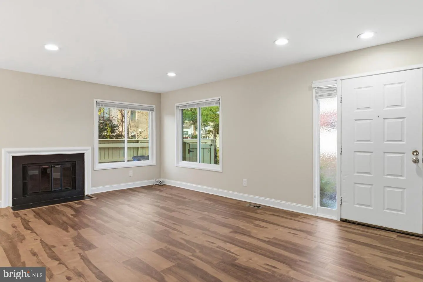 Bright living room with wood floors, fireplace, and two windows. A white front door with a glass panel is on the right.