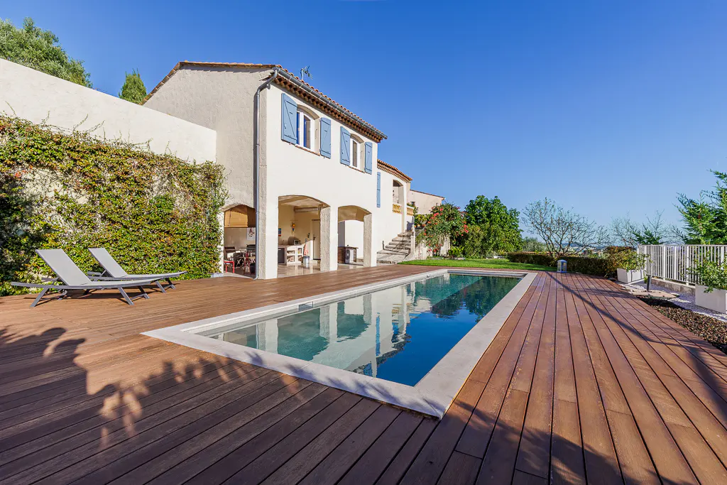 Exterior view of a white house with blue shutters, a pool, and a wooden deck under a clear blue sky.