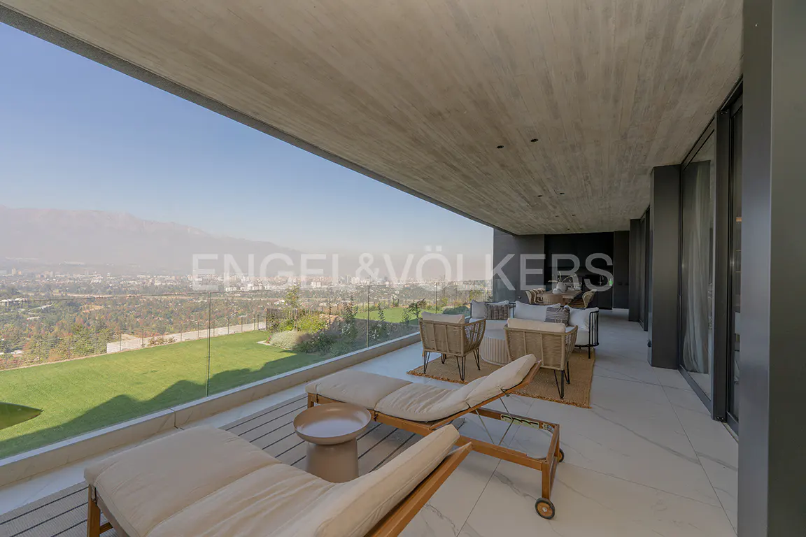 A modern patio with lounge chairs overlooks a green lawn and city view. The patio has a concrete ceiling and glass railing.