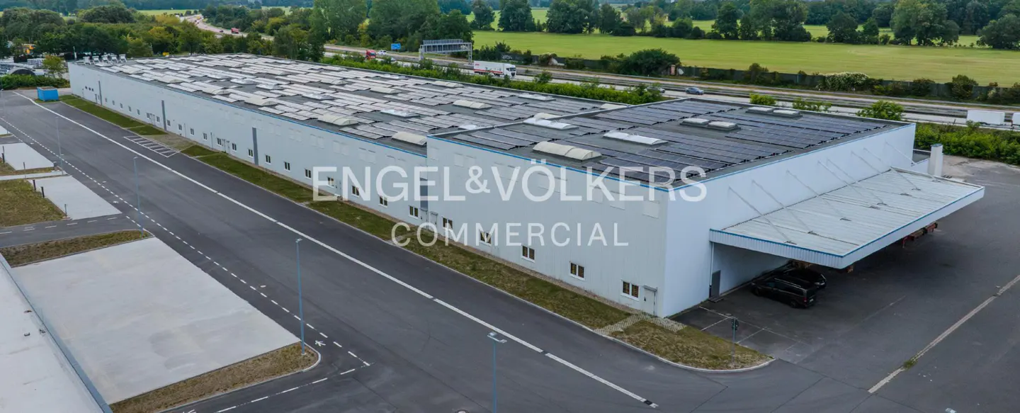 Aerial view of a long, white commercial building with solar panels on the roof, next to a highway and green fields. "Engel & Völkers Commercial" is overlaid.