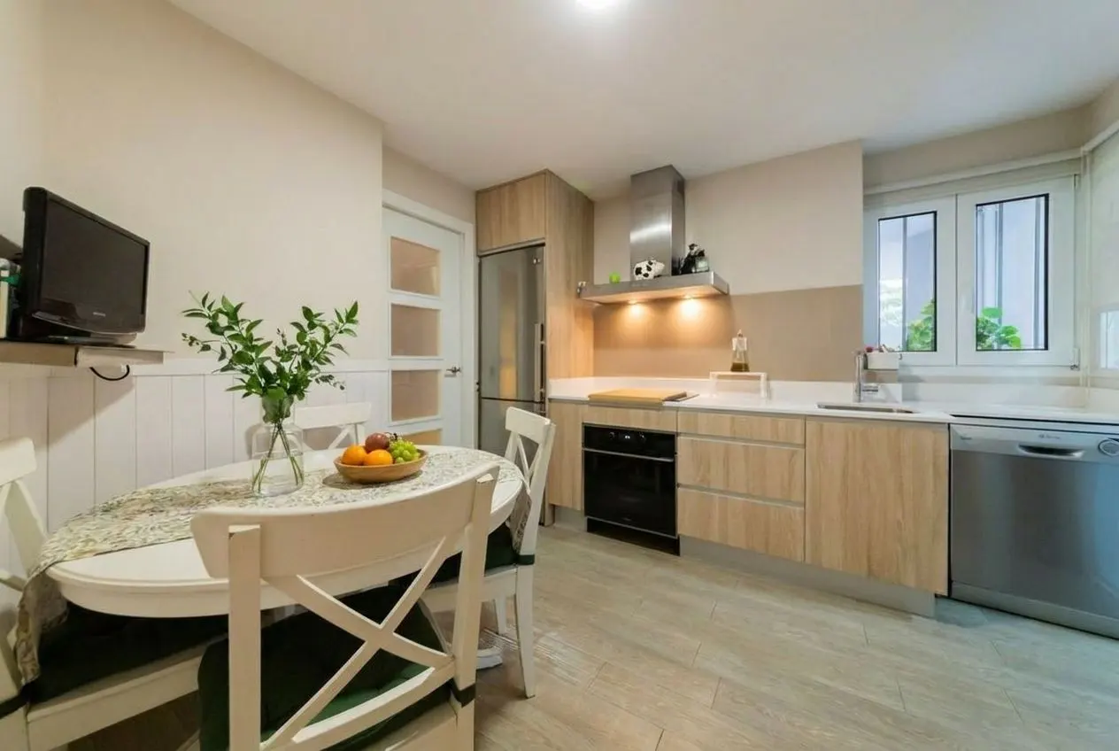 Bright kitchen with wood cabinets, stainless steel appliances, and a white table with fruit and flowers. A TV is mounted on the wall.
