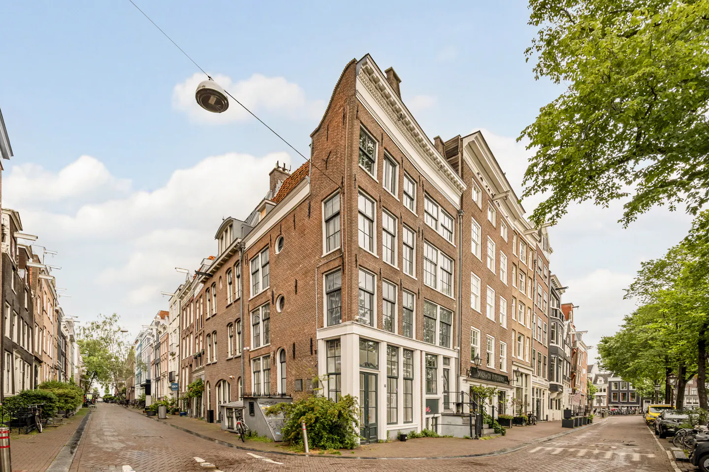 A historic red-brick corner building in Amsterdam with tall windows, situated at a street intersection on a sunny day.