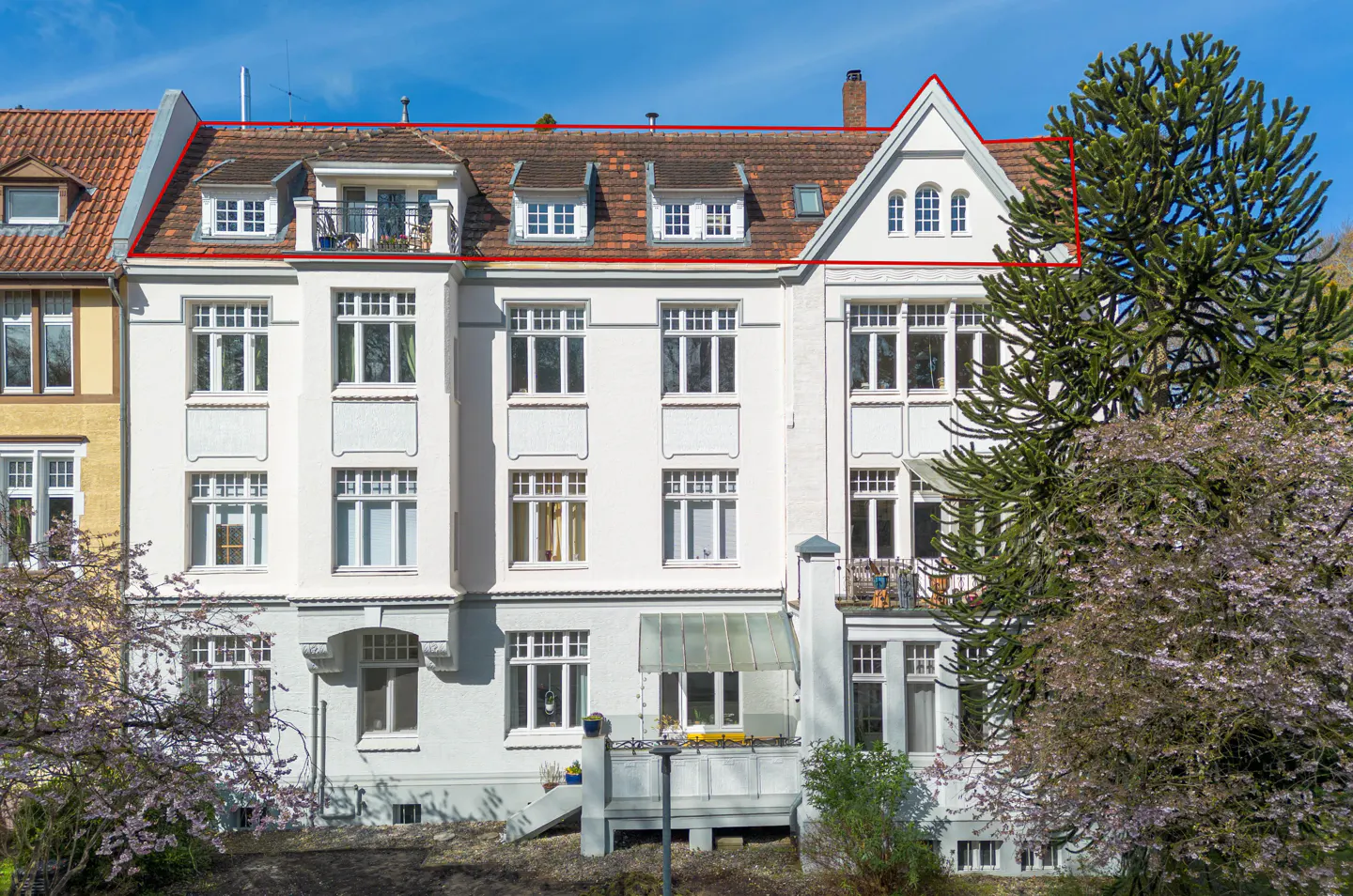Exterior view of a white, multi-story apartment building with a red-outlined roof, surrounded by trees.