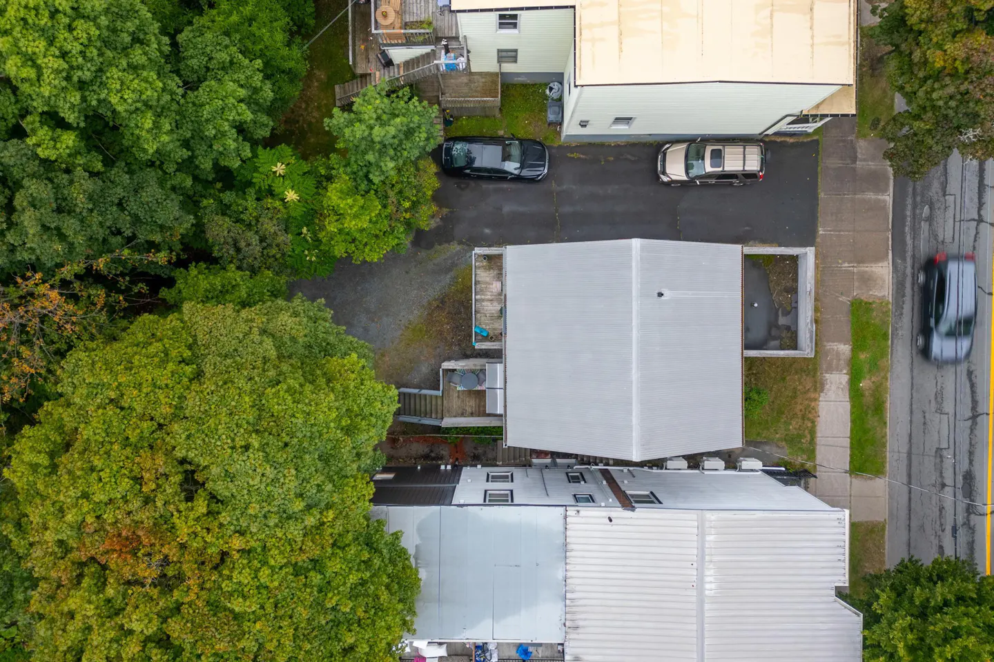 Aerial view of buildings with gray roofs, cars parked in driveways, and lush green trees surrounding the properties. A car drives on the street.