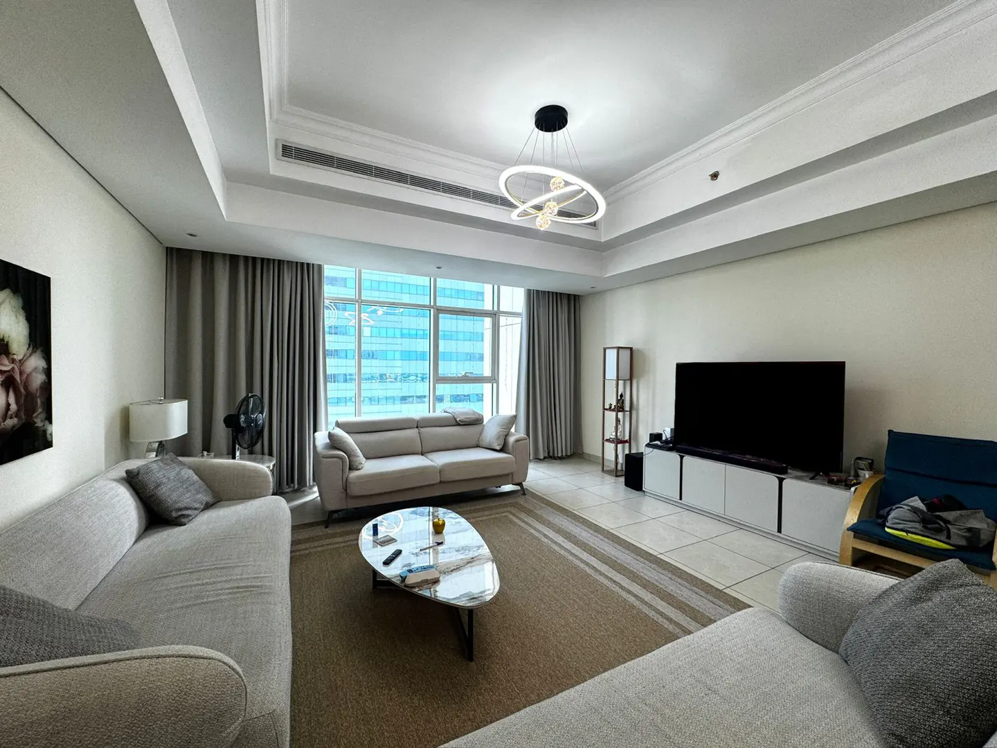 A bright living room with beige sofas, a marble coffee table, and a large window with city views.
