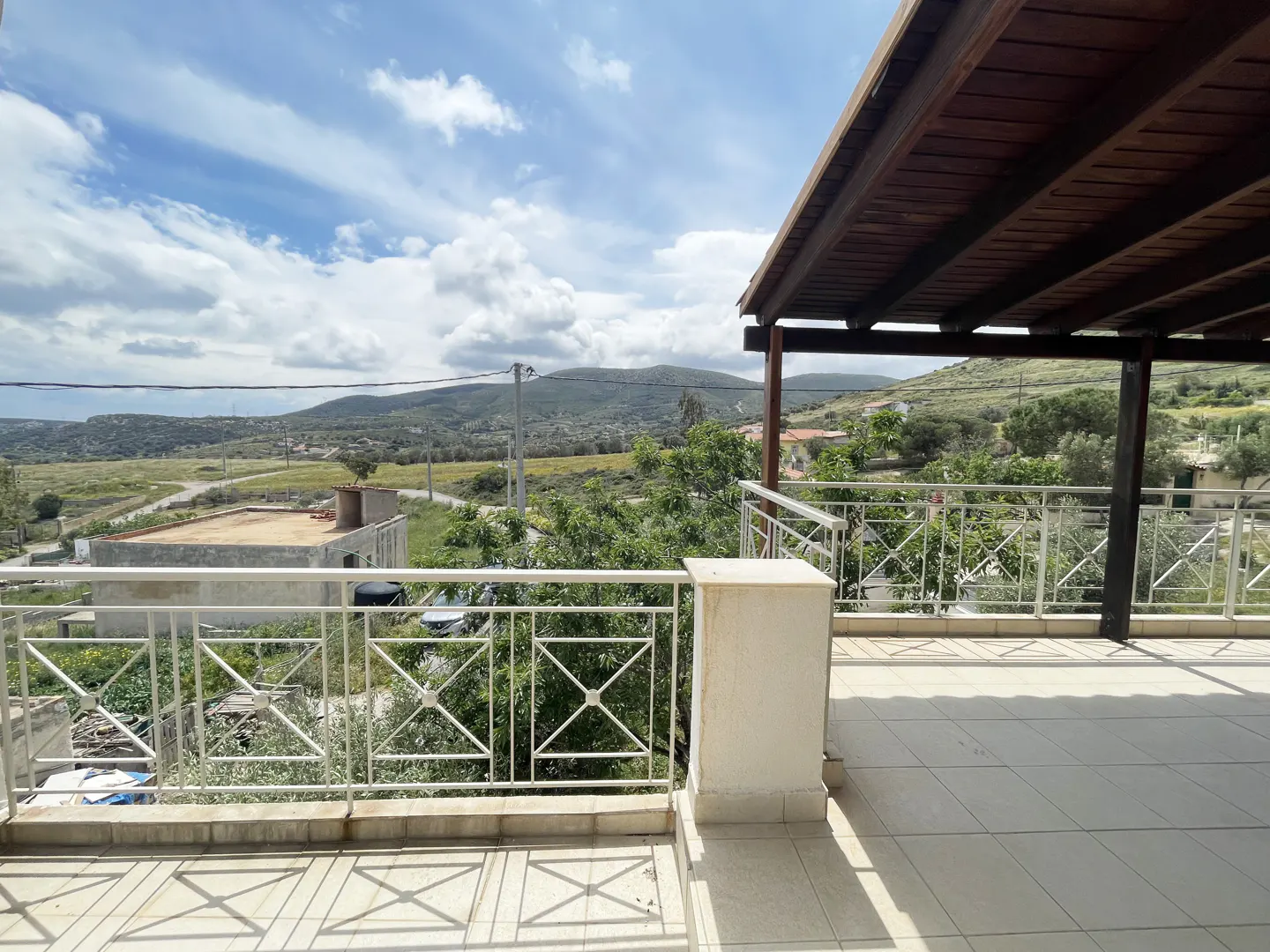 View from a tiled balcony with white railings, overlooking green hills and a cloudy sky. A wooden pergola provides shade.