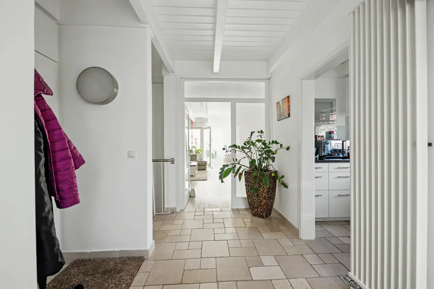 Bright hallway with white walls, tiled floor, and white wood ceiling. Coats hang near a round light fixture. A potted plant sits near an open doorway.