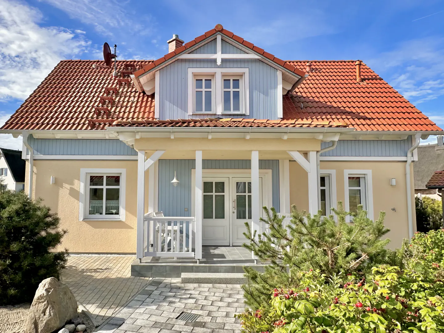 Two-story house with a red tile roof, light yellow siding, and a blue dormer. A small porch with white columns leads to double doors.