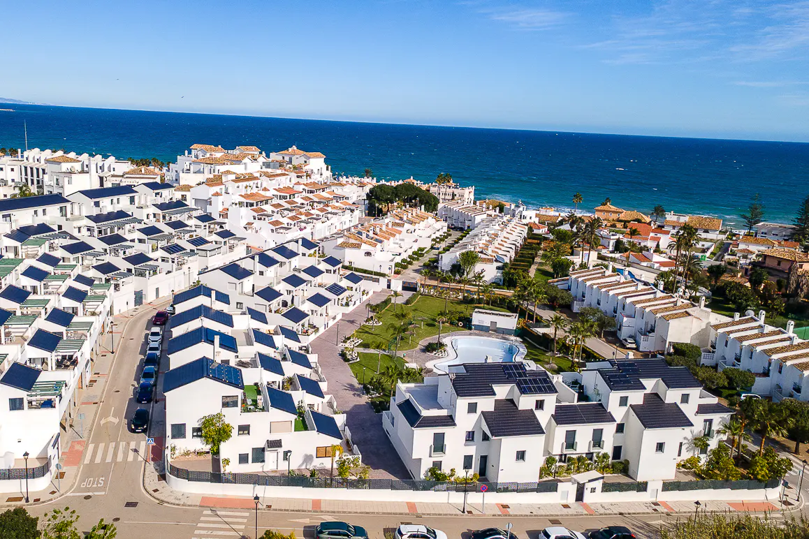 Aerial view of a coastal community with white buildings, dark roofs, and the blue ocean in the background.