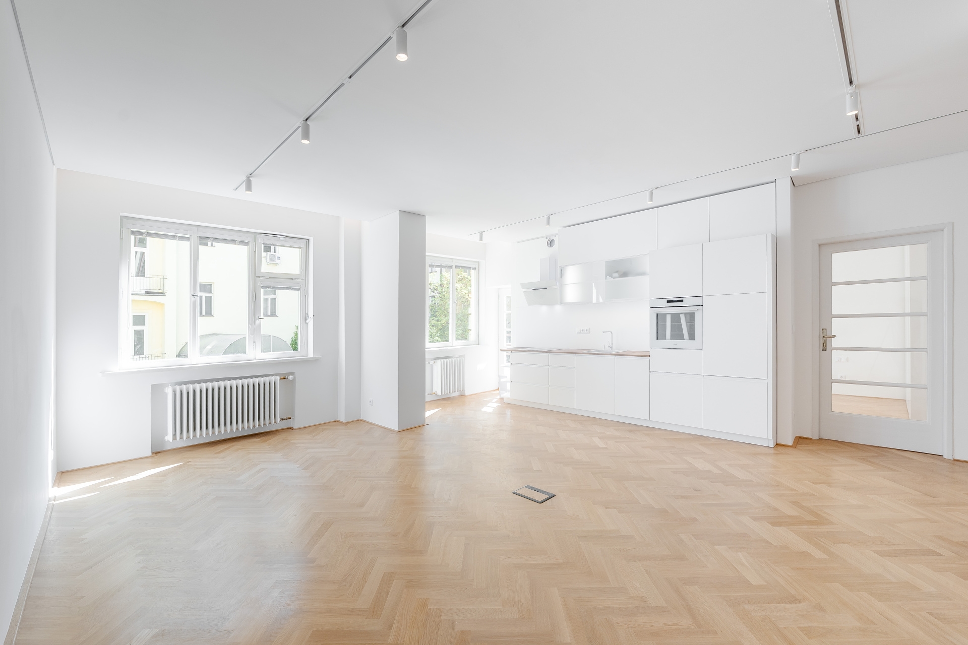Bright, minimalist open-plan kitchen and living area with white cabinets, herringbone wood floors, and track lighting. [AI-generated text]