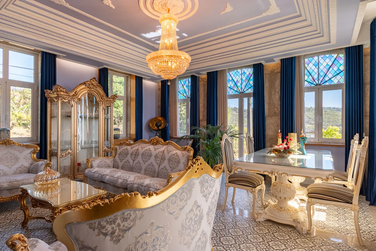 Opulent living room with gold-trimmed furniture, a crystal chandelier, and blue curtains framing windows with stained glass accents.