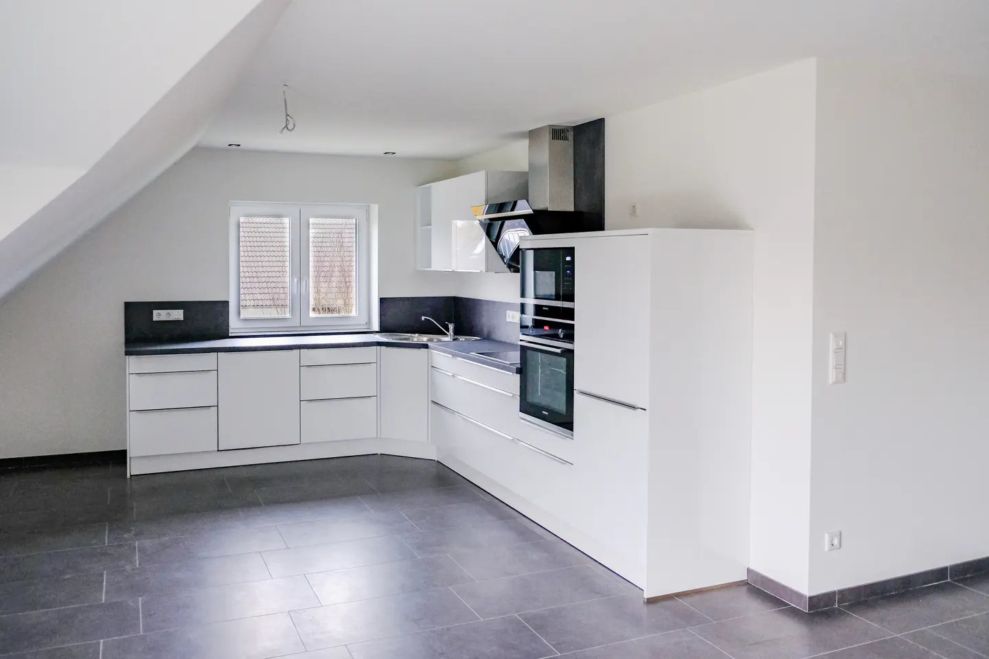 Bright, modern kitchen with white cabinets, dark countertops, and gray tile floor. Stainless steel appliances and a window add to the clean design.
