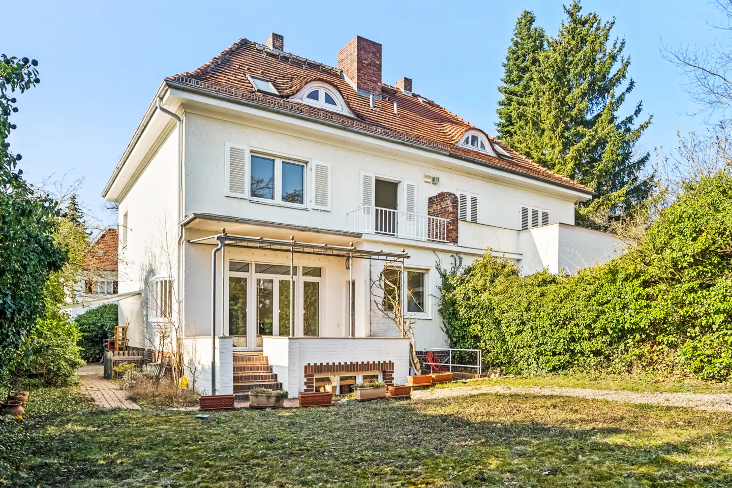 Two-story white house with a red tile roof, chimney, and a covered patio in the backyard. Green trees and bushes surround the house.