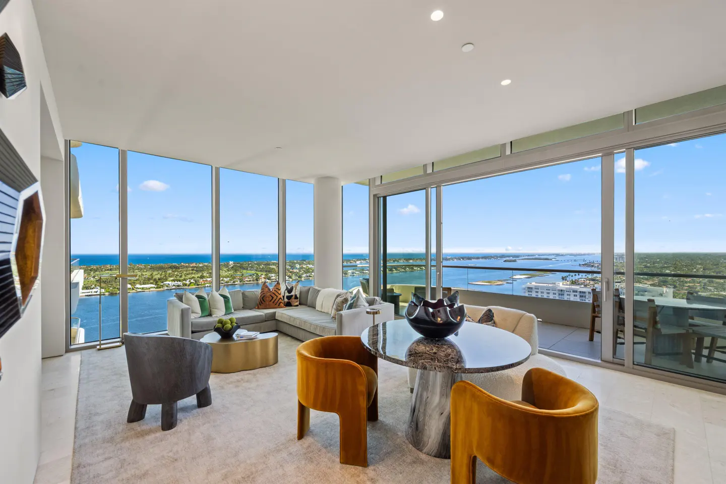 Bright, modern living room with floor-to-ceiling windows overlooking the ocean. Gray sofa, gold table, and orange chairs.