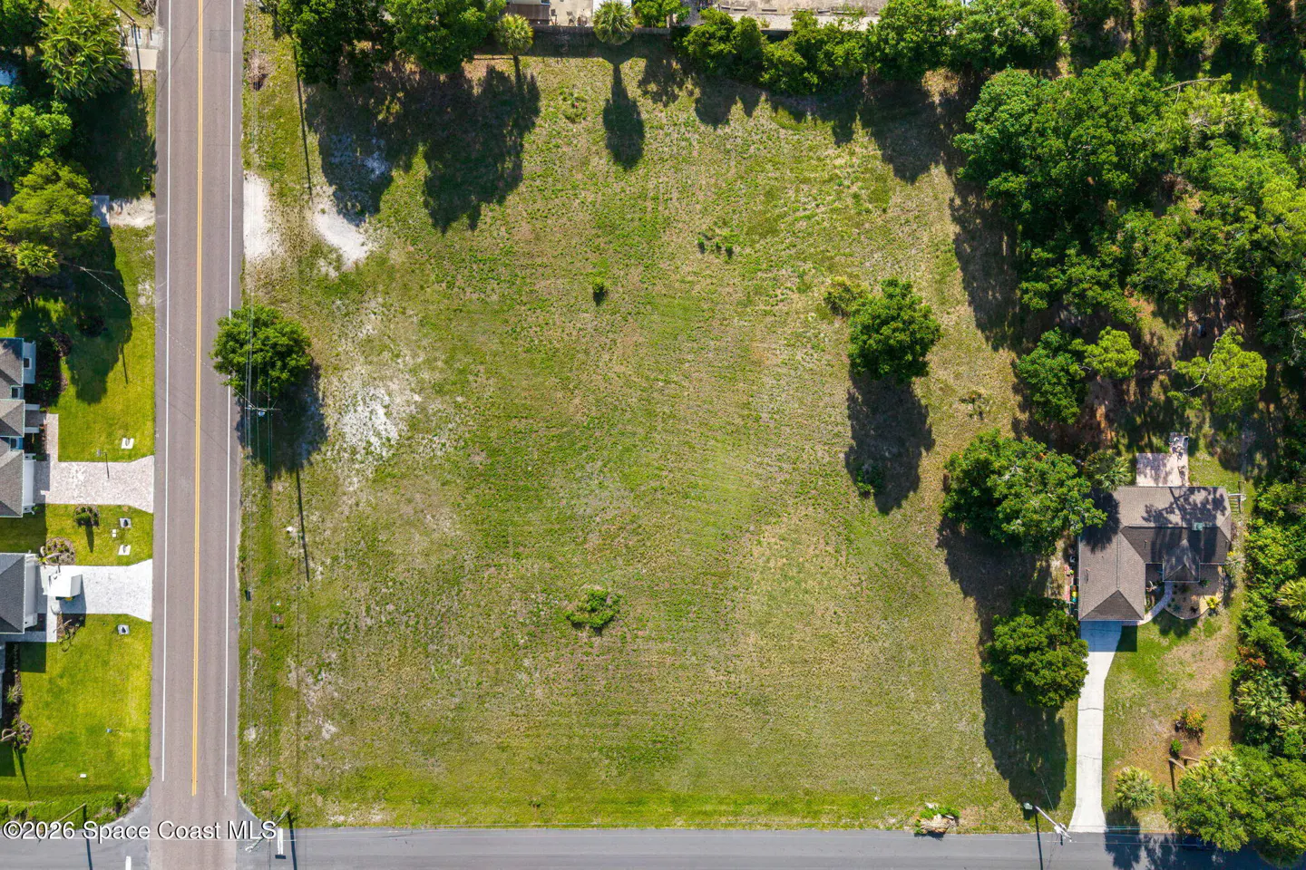 Aerial view of a vacant, grassy rectangular lot bordered by a paved road, trees, and a nearby residential home.