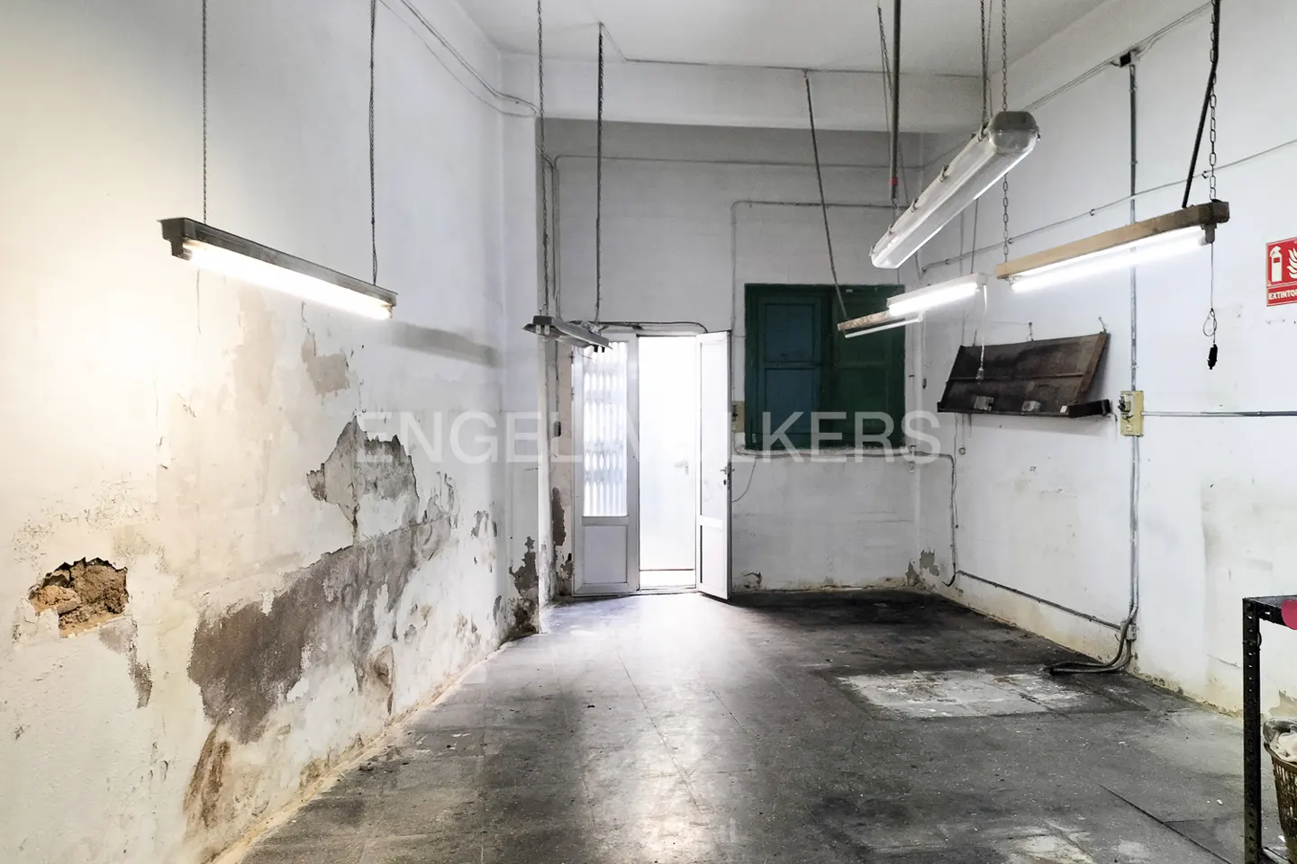 Empty room with peeling white walls, concrete floor, and fluorescent lights. A door is open at the back.