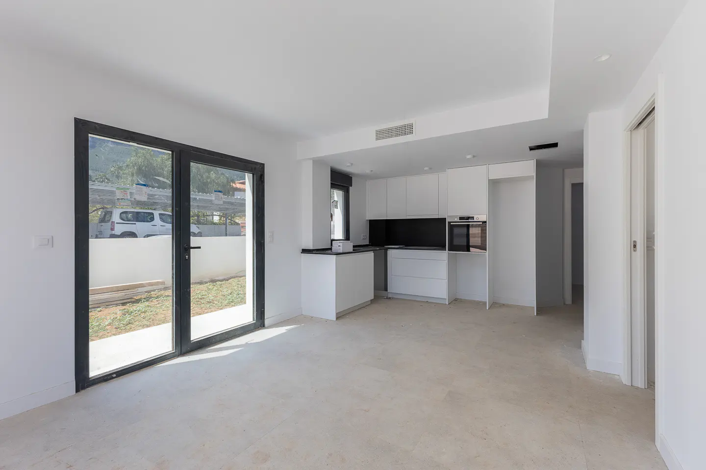 Modern minimalist kitchen with white cabinets, black backsplash, and glass doors opening to a yard with a parked car.