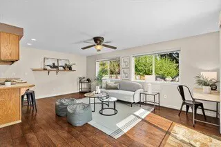 Living room with hardwood floors, a gray sofa, two beanbag chairs, and a ceiling fan. Windows overlook green trees.