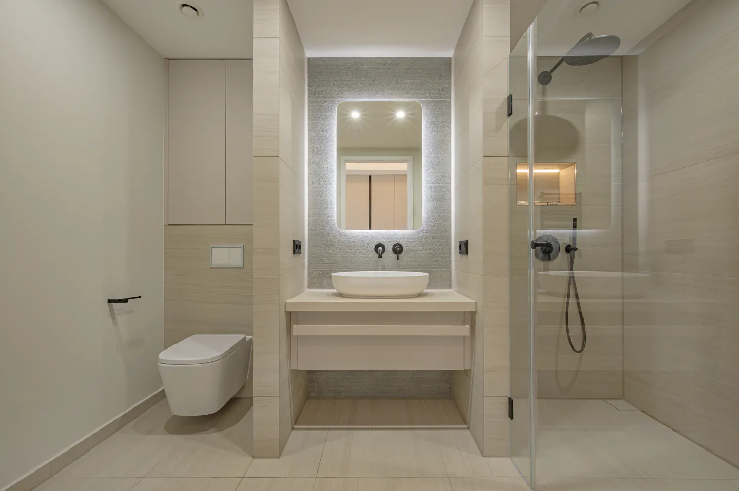 Bright bathroom with beige tile, a white toilet, and a glass shower. A vanity with a lighted mirror is centered.