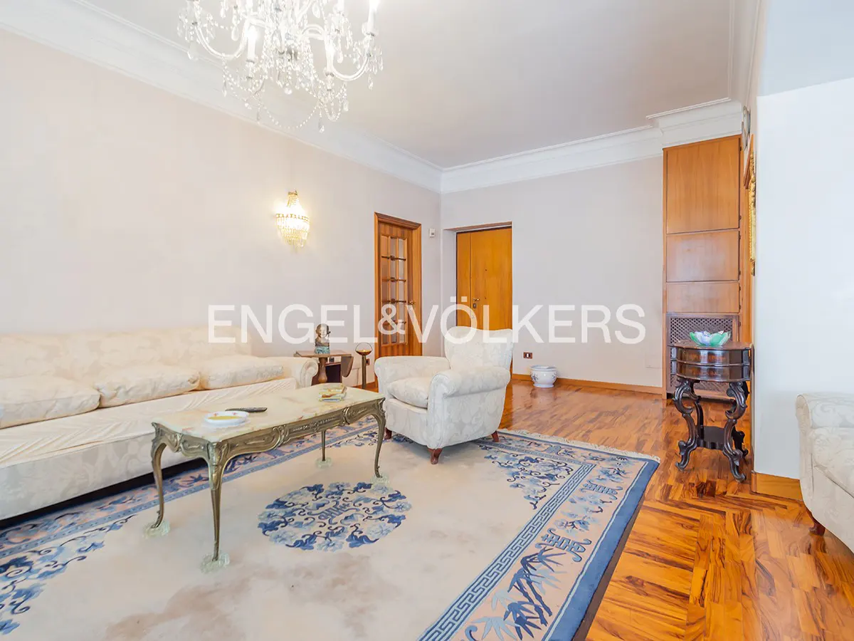 A living room with a white sofa, armchair, marble coffee table, and a blue patterned rug on a wooden floor. A chandelier hangs from the ceiling.