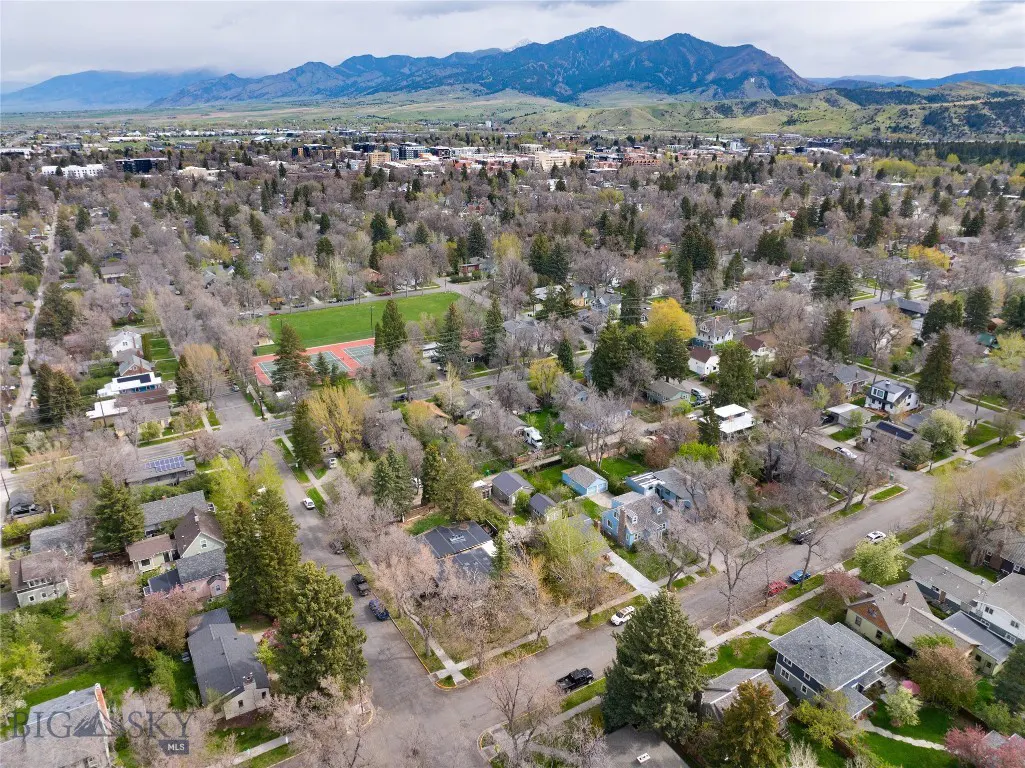 Aerial view of a residential neighborhood with houses, trees, streets, a green field, and mountains in the background.