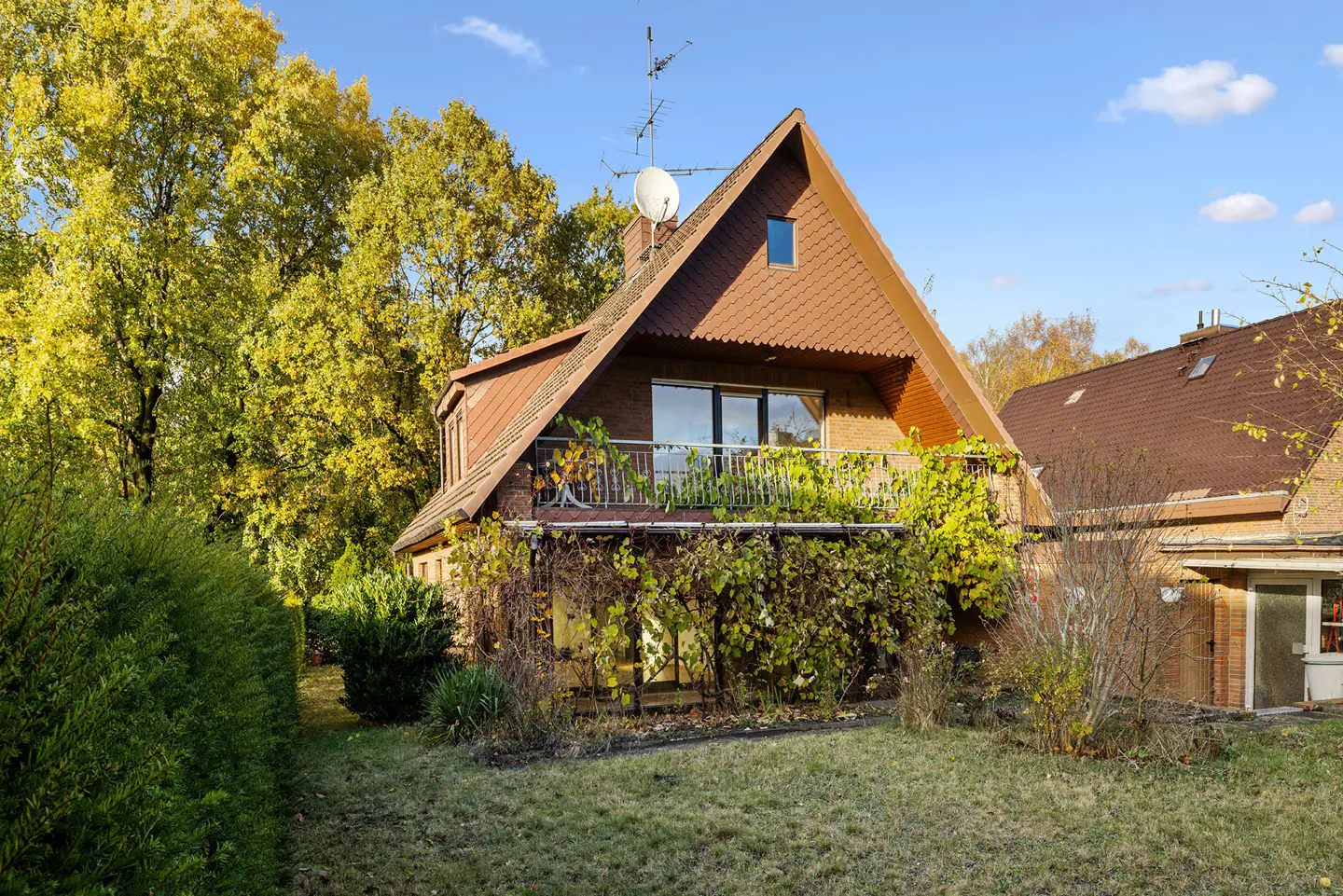 A two-story brick house with a brown roof and a balcony covered in vines. Trees with yellow leaves are in the background.