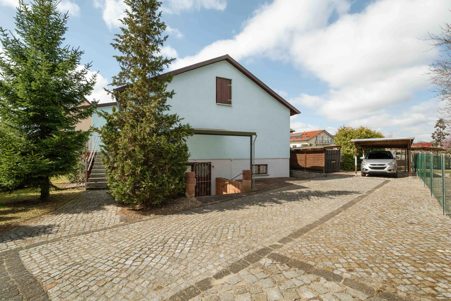 Light blue house with a cobblestone driveway, a carport with a parked car, and tall evergreen trees under a cloudy sky.