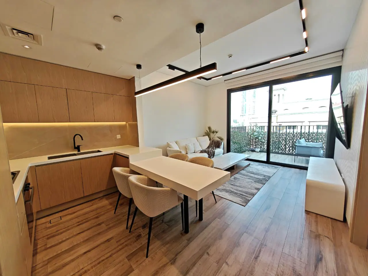 A modern apartment interior with wood floors, a kitchen with light wood cabinets, a white dining table with four chairs, and a living room with a white sofa. A balcony is visible through sliding glass doors.