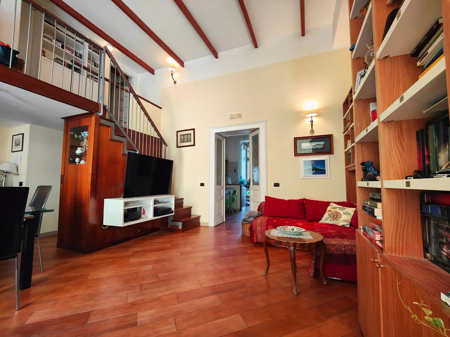 Bright living room with wood floors, red sofa, and tall bookcase. Stairs lead to a loft above a TV and cabinet. Beamed ceiling.