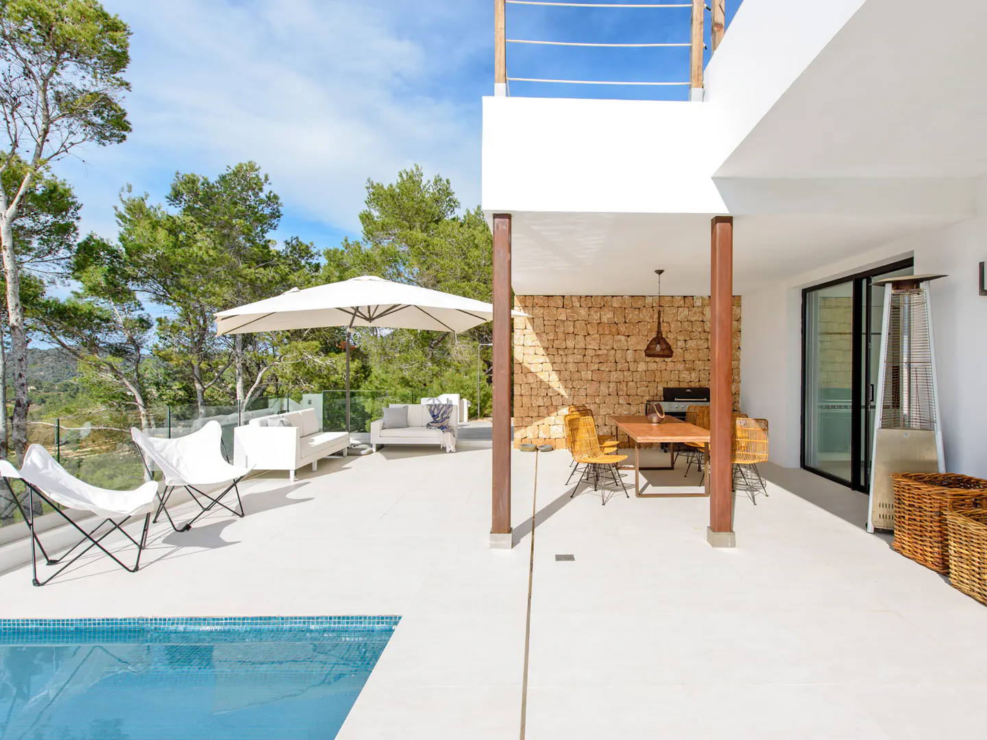 Outdoor patio with pool, white lounge chairs, and dining area with stone wall. Trees and blue sky in the background.
