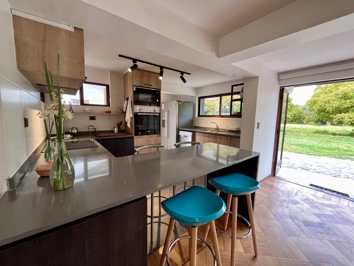 A modern kitchen with wood cabinets, gray countertops, and two turquoise bar stools at the island. An open door leads to a green lawn.