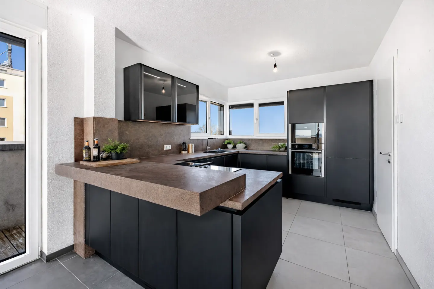 A modern kitchen with black cabinets, brown countertops, and stainless steel appliances. A balcony is visible through a glass door.
