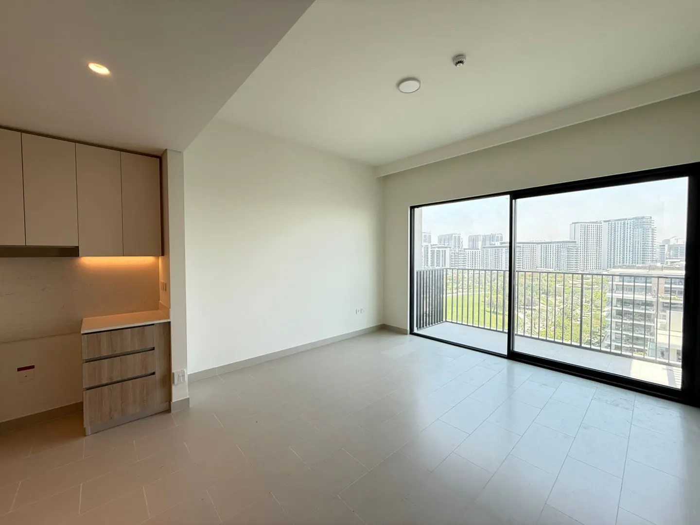 Bright, empty apartment with beige cabinets, tile floor, and sliding glass doors to a balcony overlooking a city skyline.