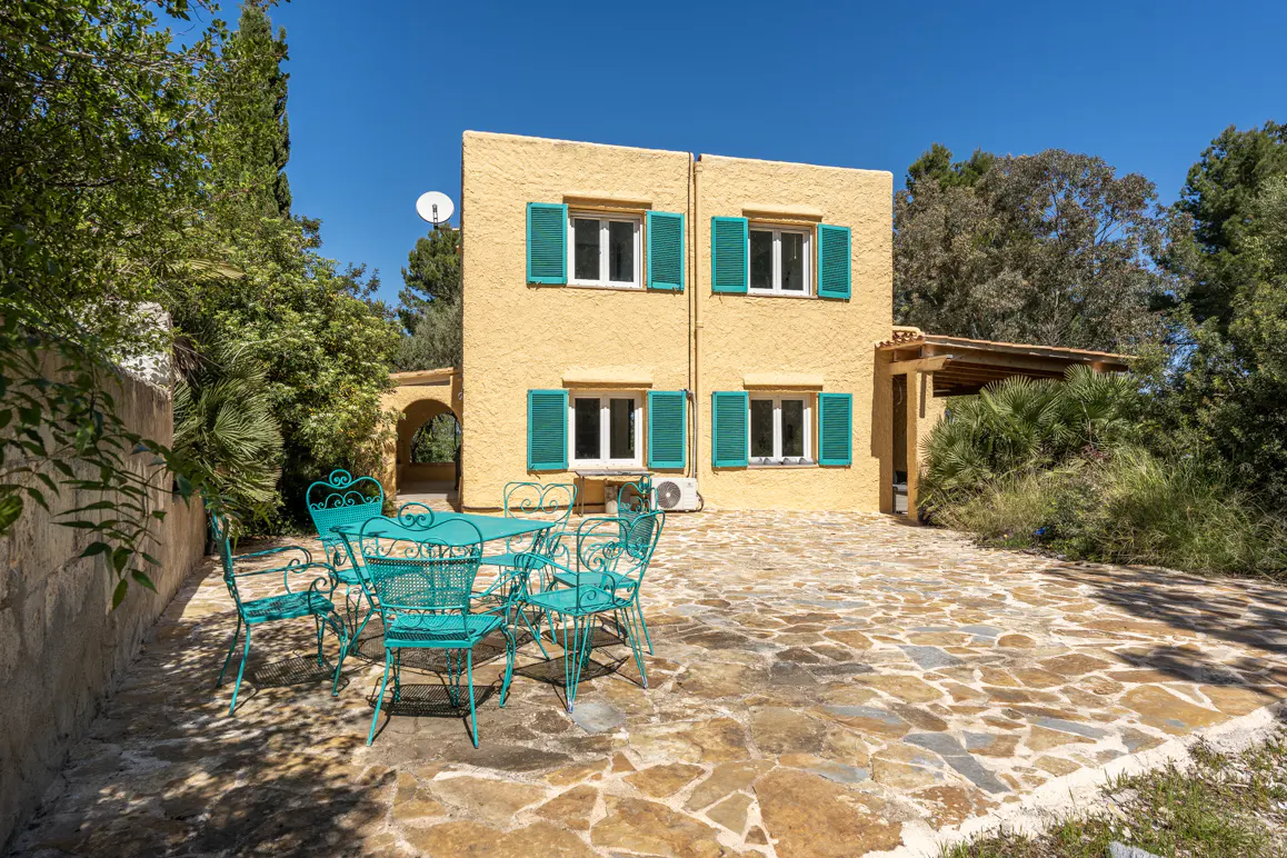 Two-story yellow house with green shutters, a stone patio, and a turquoise table and chairs.