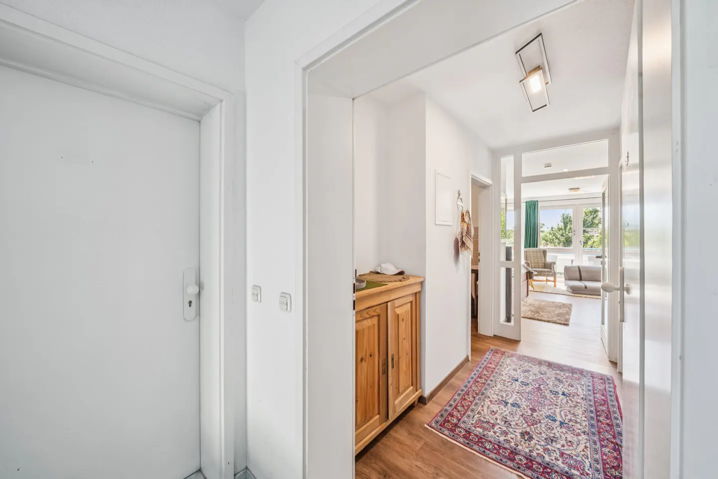 Bright hallway with white walls, a wooden cabinet, a patterned rug, and a view into a sunlit living room.