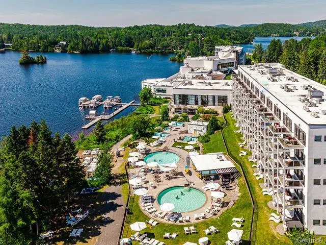 Aerial view of a lakeside resort with pools, docks, and a modern white building complex surrounded by lush green trees.
