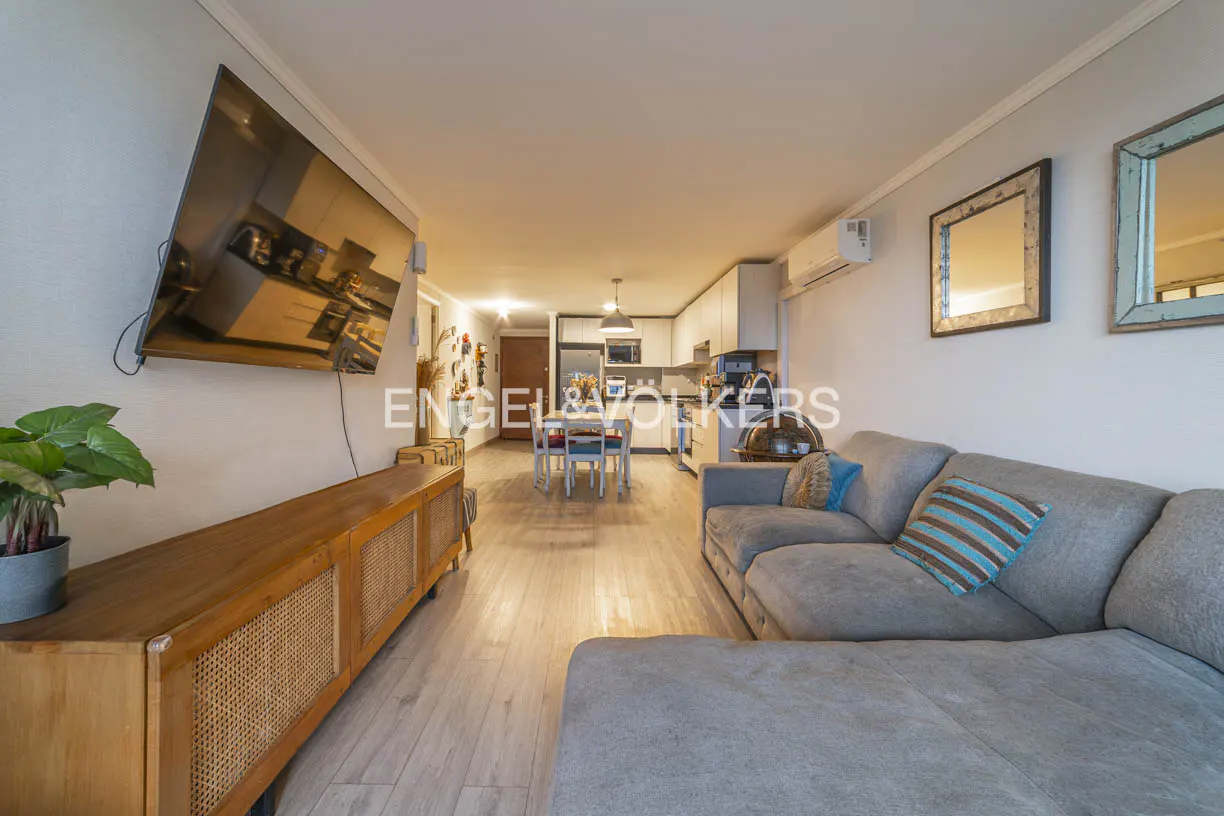 Open-plan living room with a grey sectional sofa, wooden media console, wall-mounted TV, and dining area in the background.