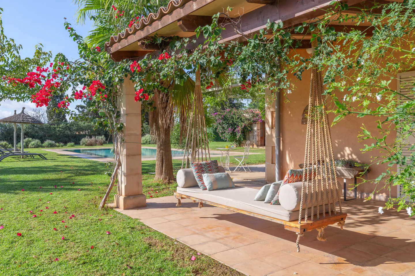 Outdoor patio with a rope swing daybed, pillows, and a pergola covered in red flowers. A pool and lawn are in the background.