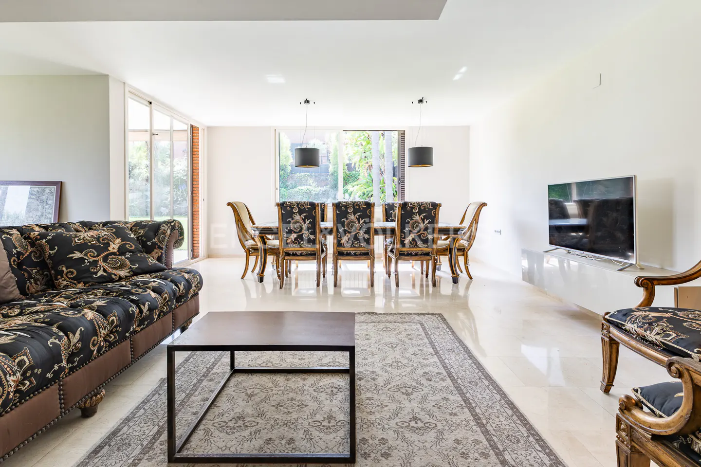 Bright living and dining area with black patterned sofa and chairs, a dark wood table, and a large TV.